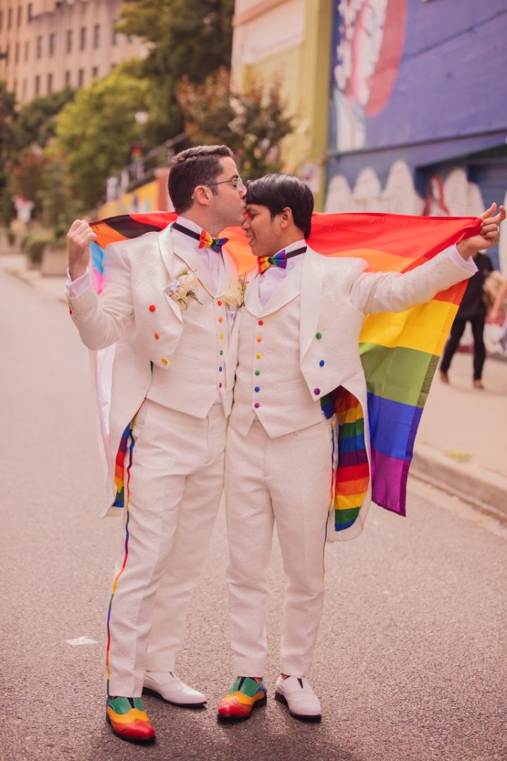 Two people wearing light tuxedos with a rainbow flag behind them